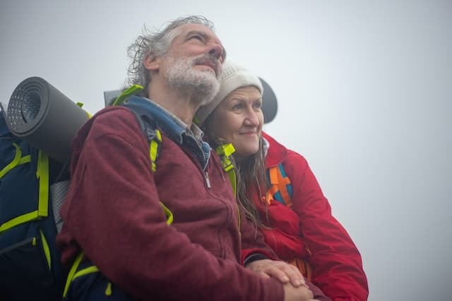 An older couple of hikers with backpacks on stand smiling in the mist.