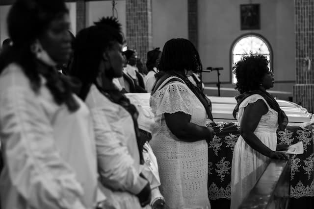 Four women dressed in white stand before a white casket at a funeral