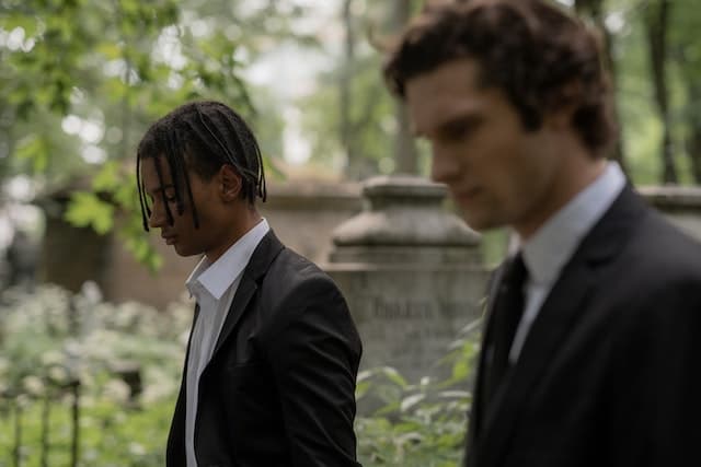Two young men in suits stand in a cemetery looking downwards.