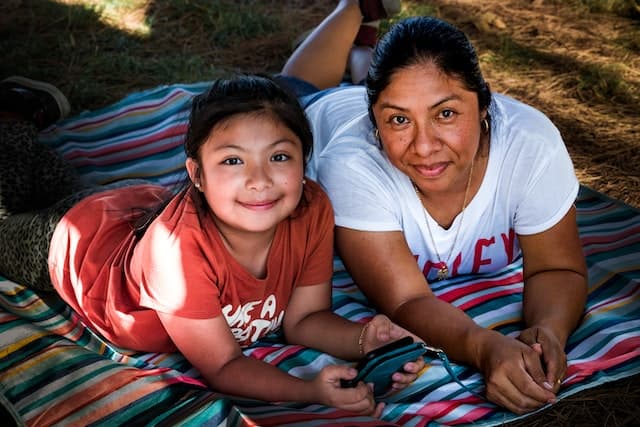 A young girl lies on a picnic rug with her mother, and both are smiling at the camera