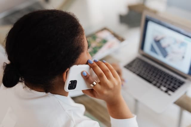 Woman dressed in white sitting at a computer talks on the phone.