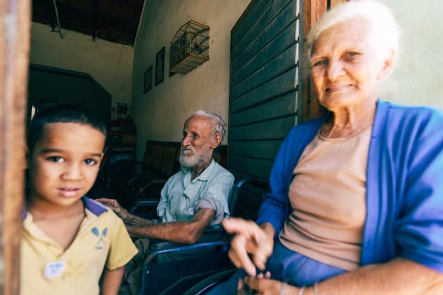 A little boy and elderly woman appear in the foreground, looking at the camera, but between them a short distance away, sits an elderly man in profile, looking at something else.