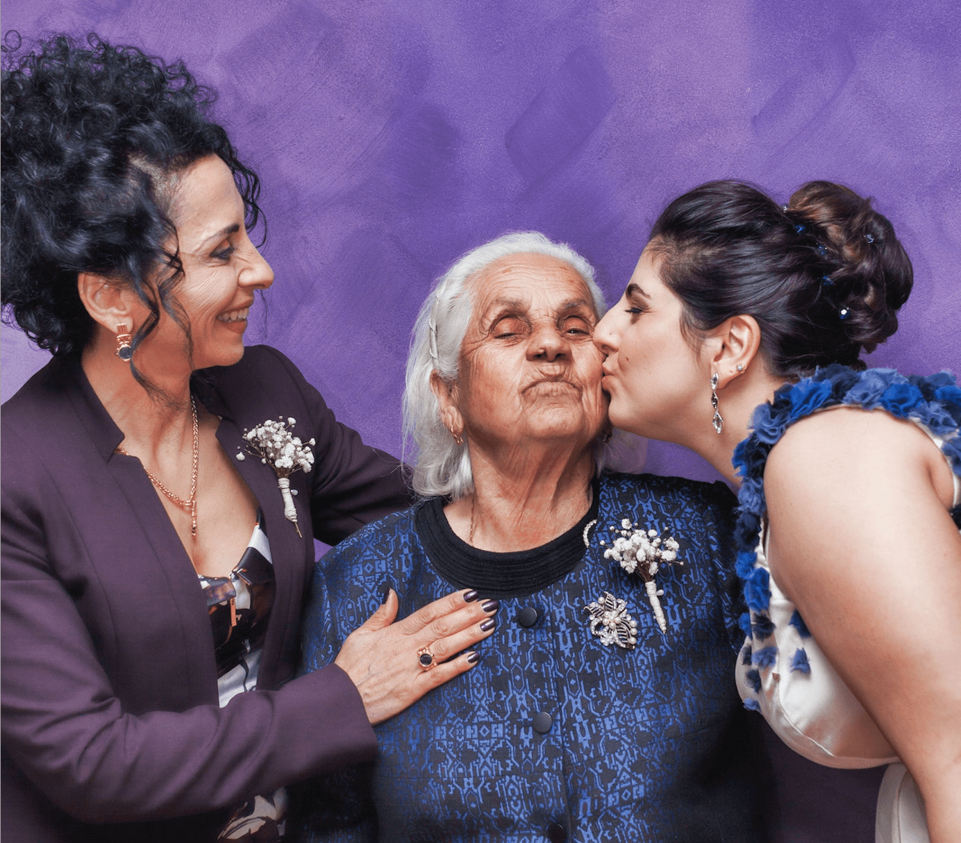 A grandma in blue is flanked by her daughter on the left, in purple, and her granddaughter on the right, who kisses her cheek.