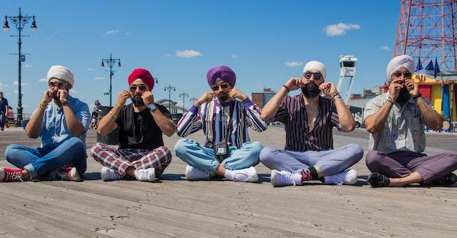Five Sikh men in colorful clothing sit in a row with their legs crossed, twirling their moustaches.