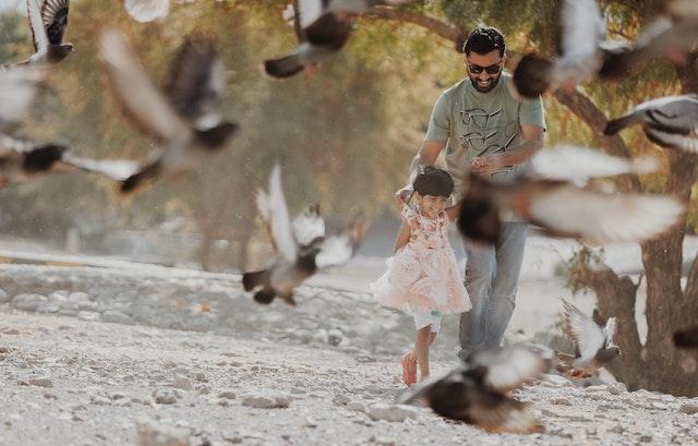 A father walks with his smiling daughter through a flock of pigeons.