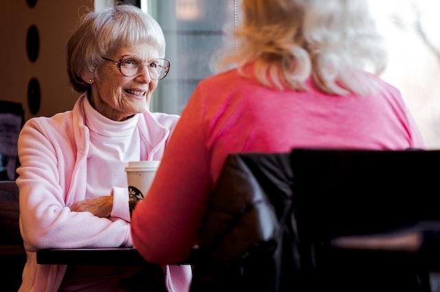 A lady with white hair and a pink jacket smiles over coffee at a woman with blonde hair, who has her back to us.