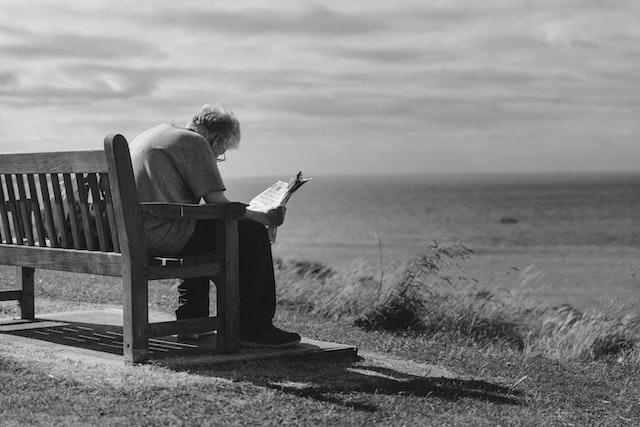 A person with greyish hair and glasses sits on a bench overlooking the ocean, and reads a newspaper.