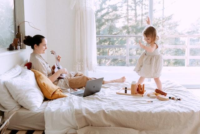 A woman sits on a bed playing ukelele to her small daughter, who dances.