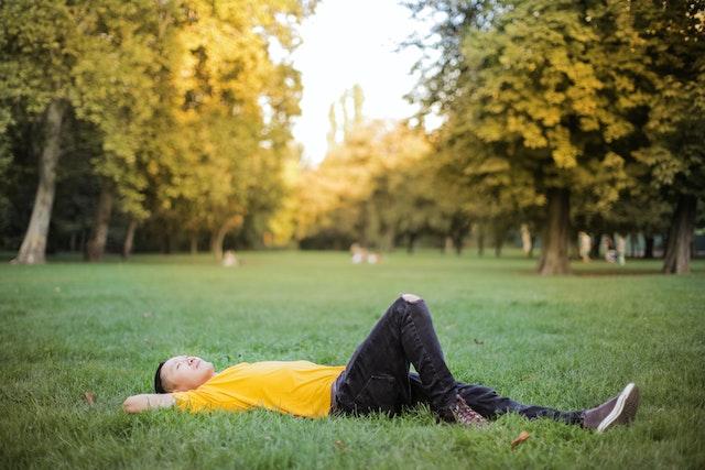 A man in a yellow t-shirt and black jeans lies in a lush, green park gazing at the sky.