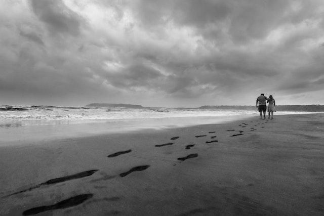 A beach overarched by a cloudy sky. To the left we see footprints left by the two people who appear further along the waterline to the right.