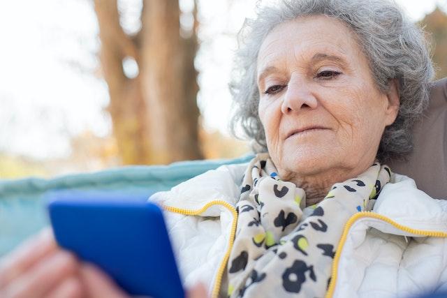 An older woman seated on an outdoor sofa reads on her phone.